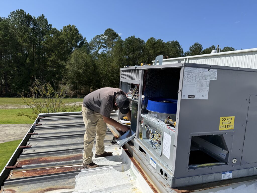Technician repairing an HVAC unit on a rooftop under a clear blue sky, surrounded by trees.